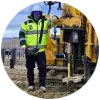 A person in a high-visibility jacket operates construction equipment at a work site with metal posts in the background.