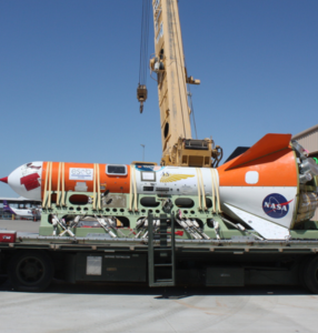 Aerospace vehicle on a flatbed trailer, featuring orange and white colors with NASA and other logos. A crane is positioned overhead under a clear blue sky.