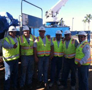 Six construction workers in safety vests and hard hats stand side by side at a work site with machinery in the background.