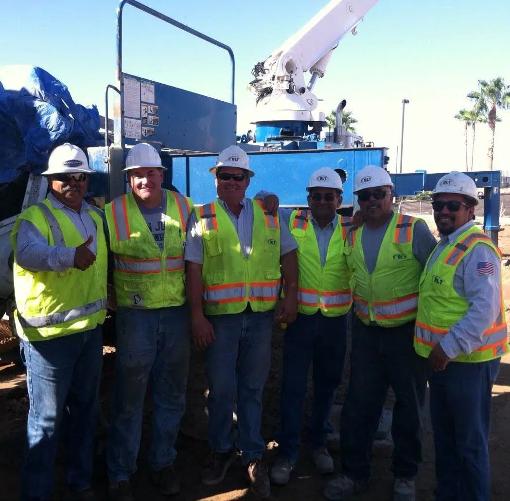 Six construction workers in safety vests and hard hats stand side by side at a work site with machinery in the background.