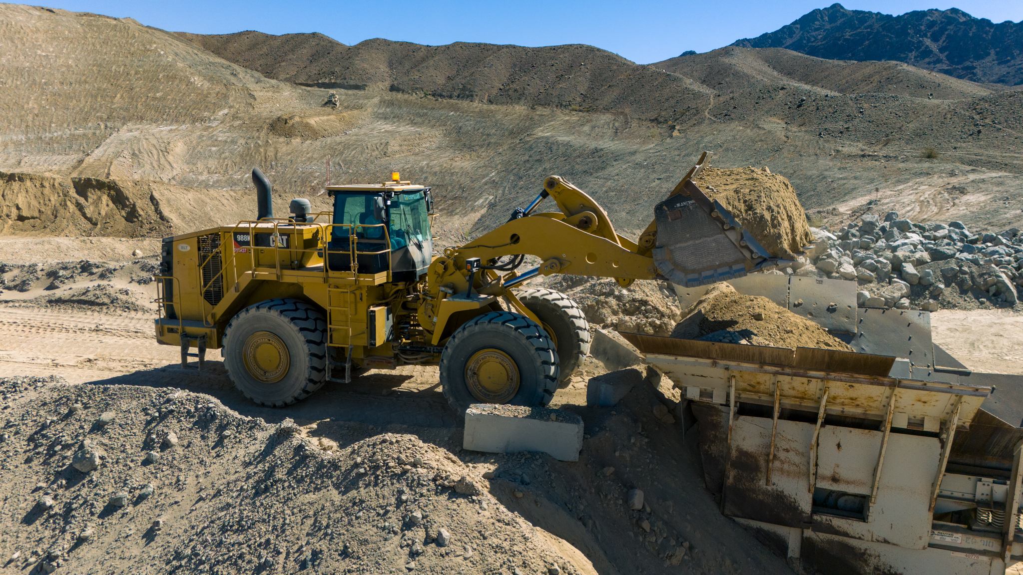 A yellow bulldozer moves dirt at a mining site, with rocky hills in the background under a clear blue sky.