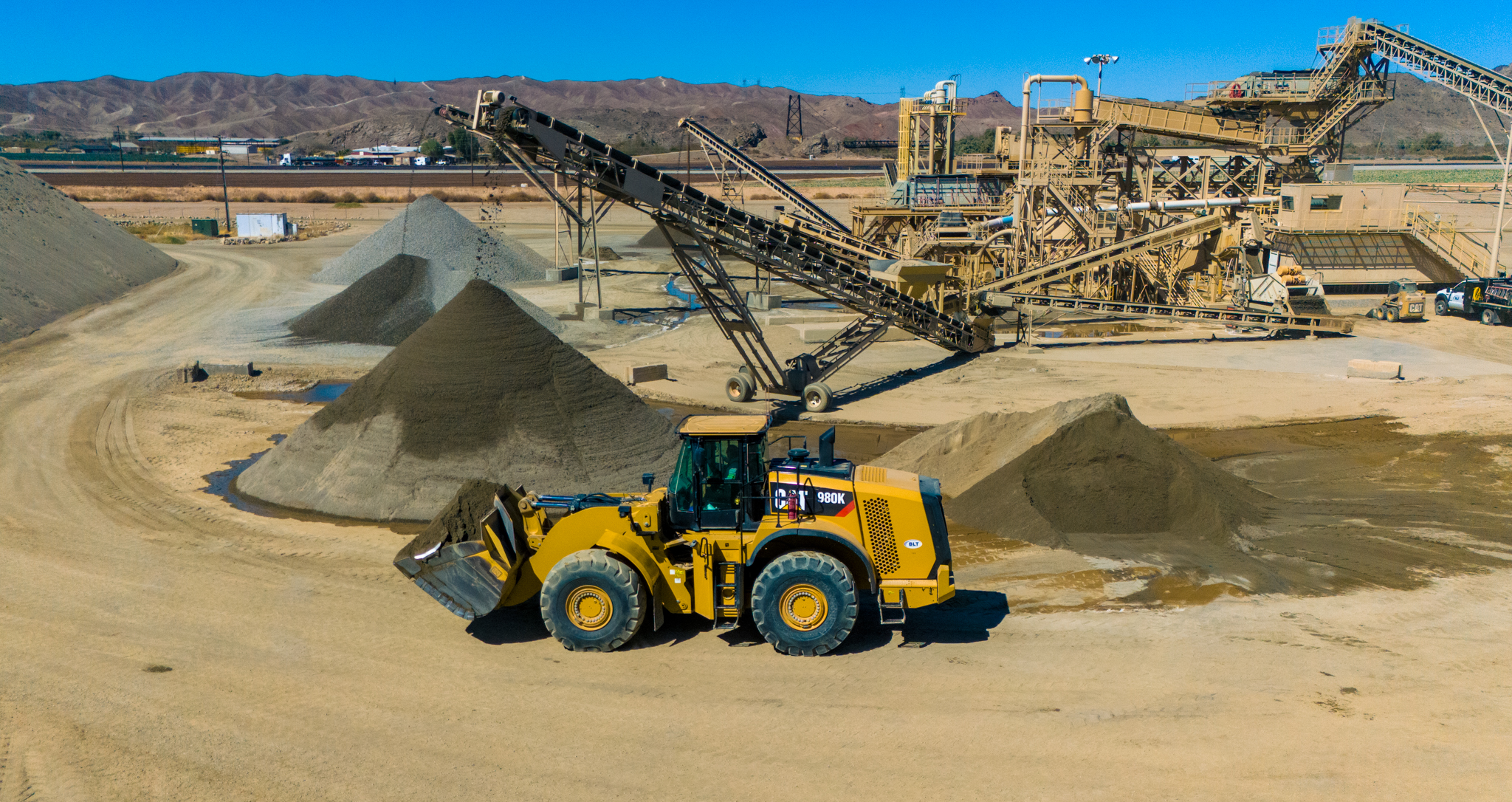 A yellow bulldozer moves sand in a quarry with conveyor belts and piles of gravel under a clear blue sky.