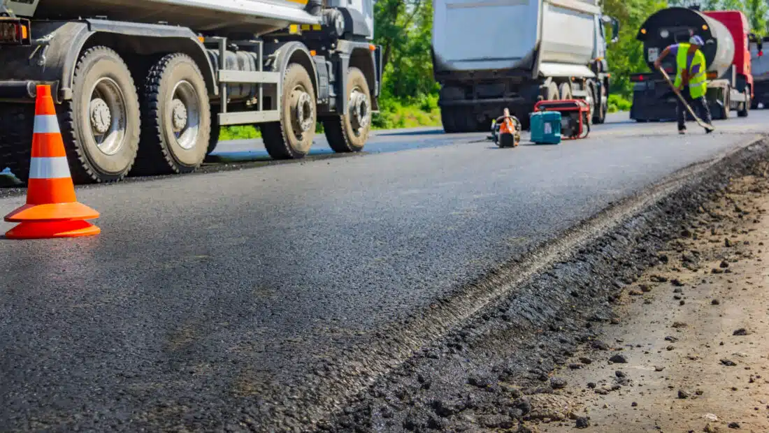 Freshly paved road with construction vehicles and workers in safety vests; traffic cones and equipment are visible along the edge of the new asphalt surface.