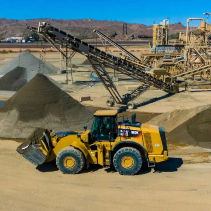 A yellow front-end loader transports dirt near large gravel piles at an industrial mining site with conveyor belts and equipment in the background.