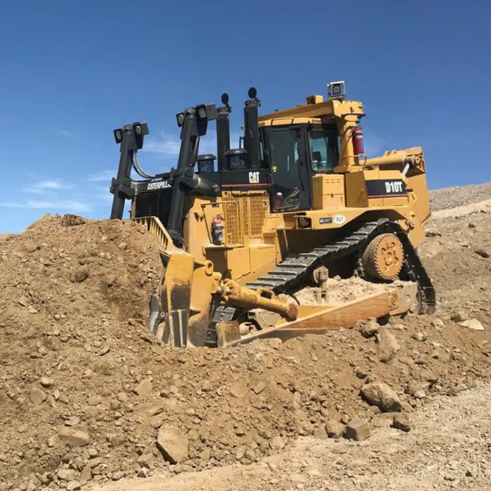 A yellow Caterpillar bulldozer moves a large pile of dirt at a construction or mining site under a clear blue sky.