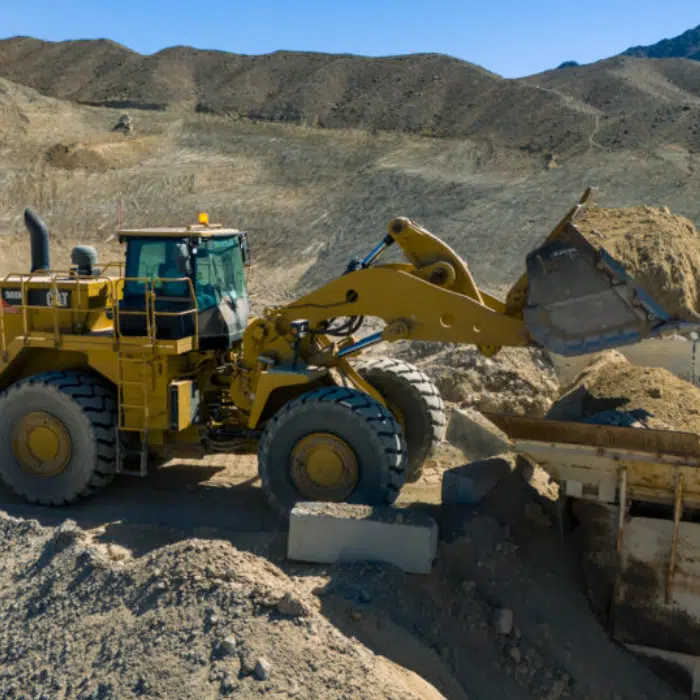 A yellow front-end loader moves a pile of dirt at a construction site with hills in the background under a clear sky.