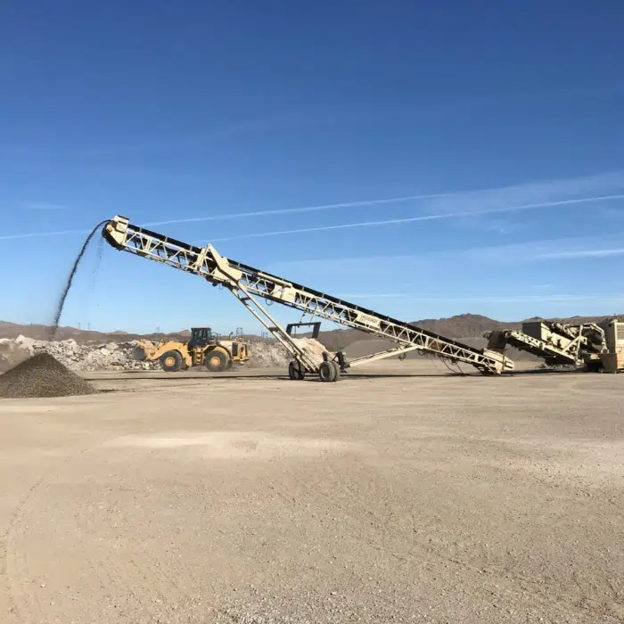 A conveyor belt deposits gravel onto a pile at an open construction or mining site, with heavy machinery in the background under a clear blue sky.