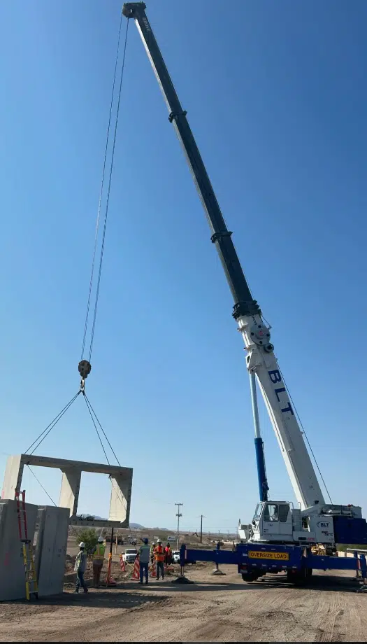 A large crane lifts a concrete structure while several construction workers stand nearby on a sunny day at a worksite.
