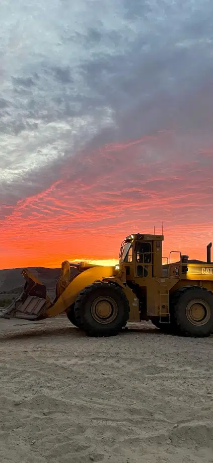 A yellow Caterpillar front loader is parked on dirt ground at sunset, with a dramatic red and orange sky in the background.